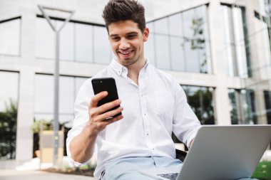 Açık havada dizüstü bilgisayar ve cep telefonu kullanarak oturan bir yakışıklı genç iş adamı görüntüsü.