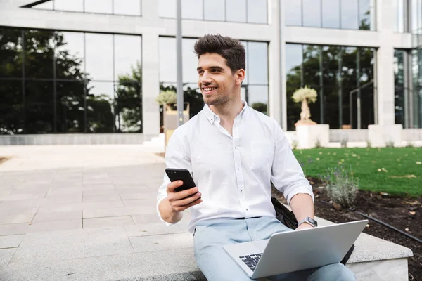 ラップトップ コンピューターや携帯電話を使用した屋外に座っているハンサムな若いビジネスマンのイメージ — ストック写真