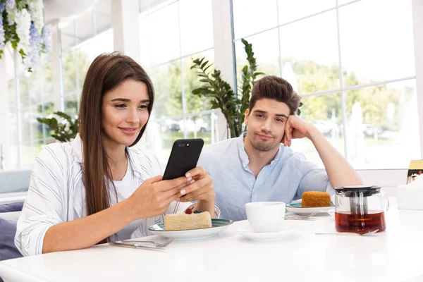 Sorrindo Jovem Sentado Mesa Café Enquanto Sua Namorada Usando Telefone — Fotografia de Stock