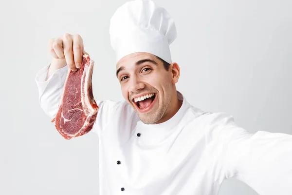 愉快的厨师厨师穿着制服 显示生牛排隔离在白色的背景 采取自拍 — 图库照片