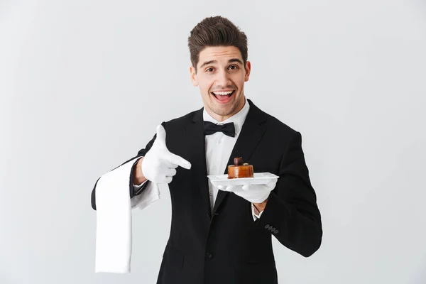 英俊的年轻服务员在燕尾服与碗举行板与蛋糕查出在白色背景 — 图库照片