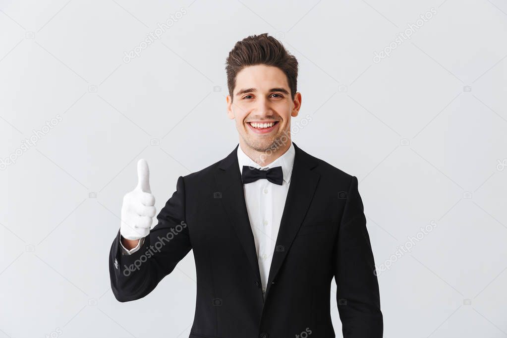 Portrait of a handsome young man waiter wearing tuxedo and gloves standing isolated over white background, showing thumbs up