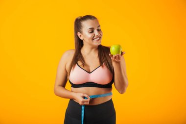 Beline metre ile ölçme ve elma izole sarı arka plan üzerinde tutan sportif sutyen fitness tombul kadın portresi