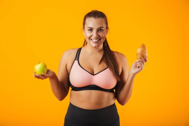 Elma ve kruvasan izole sarı arka plan üzerinde her iki elinde tutan eşofman gülümseyen tombul kadın görüntüsünü