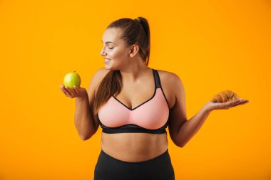 Elma ve kruvasan izole sarı arka plan üzerinde her iki elinde tutan eşofman genç tombul kadın görüntüsünü