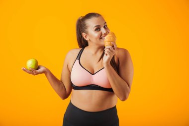 Elma tutan ve sarı arka plan üzerinde izole kruvasan yemek eşofman beyaz tombul kadın görüntüsünü