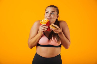 Sandviç ve patates kızartması sarı arka plan üzerinde izole yeme eşofman neşeli tombul kadın görüntüsünü