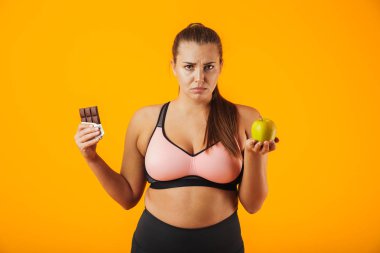 İçinde çikolata ve elma izole sarı arka plan üzerinde her iki elinde tutan eşofman memnun kilolu kadın resim