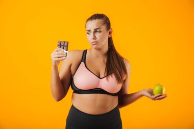 İçinde çikolata ve elma izole sarı arka plan üzerinde her iki elinde tutan eşofman üzgün tombul kadın resim