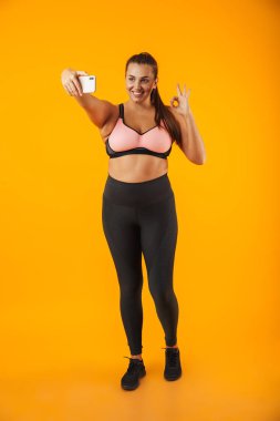 Sportif sutyen selfie fotoğrafta üstünde hareket eden telefon içinde neşeli tombul kadın portresi tam uzunlukta sarı arka plan üzerinde izole