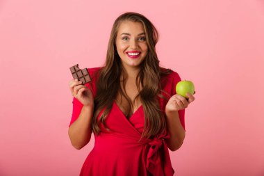Görüntü güzel bir aç genç kadının çikolata ve elma tutan pembe duvar arka plan üzerinde izole.