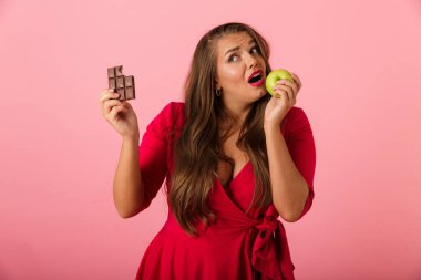 Görüntü güzel bir aç genç kadının çikolata ve elma tutan pembe duvar arka plan üzerinde izole.