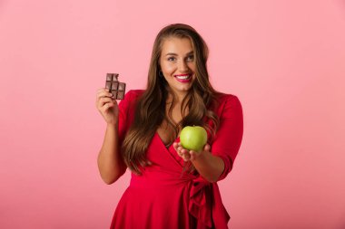 Görüntü güzel bir aç genç kadının çikolata ve elma tutan pembe duvar arka plan üzerinde izole.