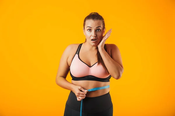 兴奋的胖乎乎的女人在运动胸罩测量她的腰与米查出在黄色背景的肖像 — 图库照片
