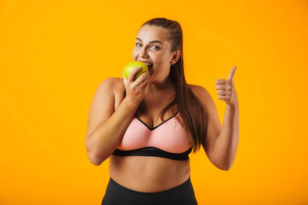 成年胖乎乎的妇女在运动胸罩吃苹果的肖像查出在黄色背景 — 图库照片