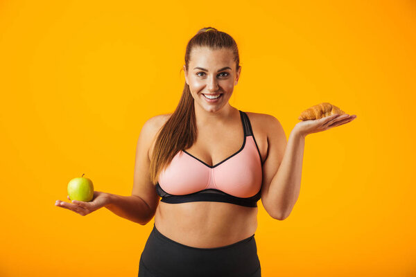 Image of european chubby woman in tracksuit holding apple and croissant in both hands isolated over yellow background