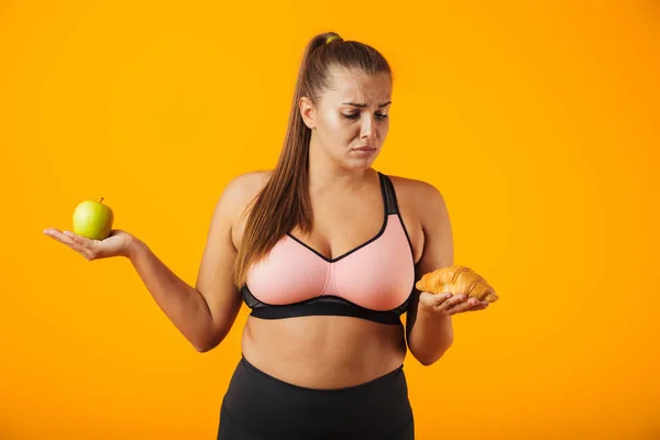 Image of displeased chubby woman in tracksuit holding apple and croissant in both hands isolated over yellow background