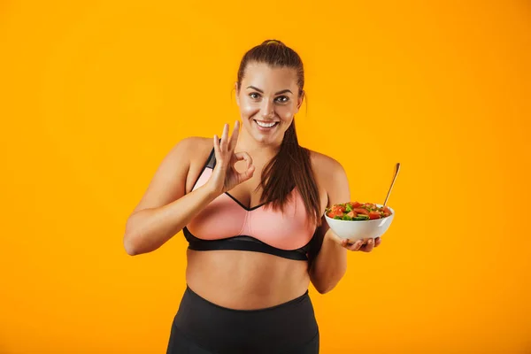 Image of cheerful chubby woman in tracksuit holding plate with green salad isolated over yellow background