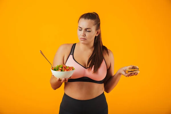 Portrait of an upset overweight fitness woman wearing sports clothing standing isolated over yellow background, holding bowl with salad and a burger