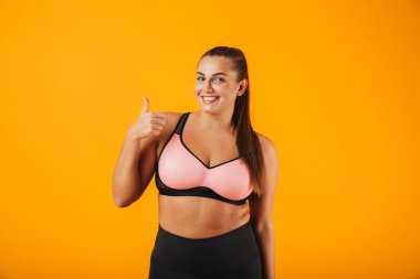 Spor giyim duran sarı arka plan üzerinde izole giyen bir neşeli kilolu fitness kadın portresi başparmak yukarı