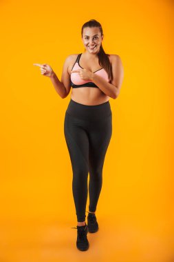 Kilolu sporcumuz gülümseyen ve bir kenara izole sarı arka plan üzerinde parmak işaret sportif sütyen tam uzunlukta portresi