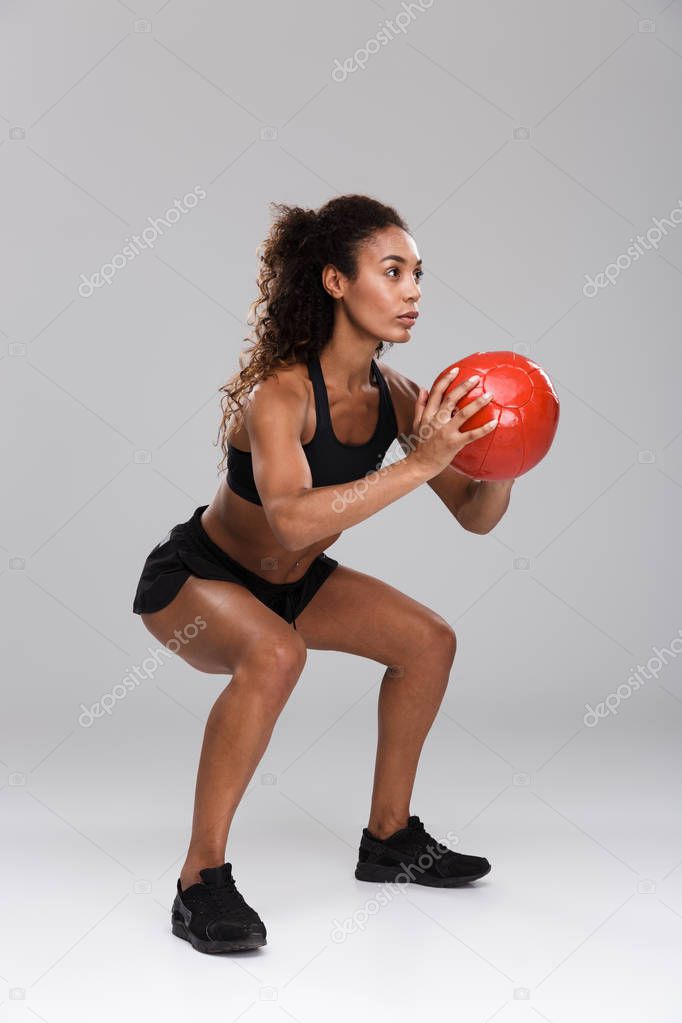 Beautiful african sportswoman isolated over gray background, doing exercises with a heavy ball, squatting