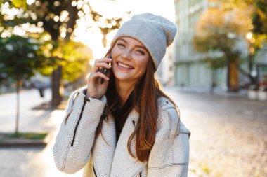 Açık havada giyen şapka cep telefonuyla konuşan yürüyen bir neşeli güzel kızıl saçlı kadın görüntüsünü.