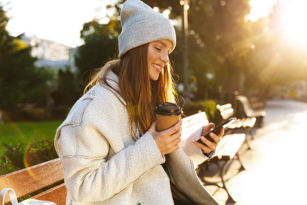 秋のコートとベンチ外に座って コーヒーを飲みながら 携帯電話を使用しての帽子に身を包んだ美しい若い女性 — ストック写真