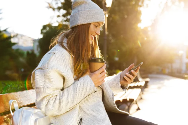 秋のコートとベンチ外に座って コーヒーを飲みながら 携帯電話を使用しての帽子に身を包んだ美しい若い女性 — ストック写真