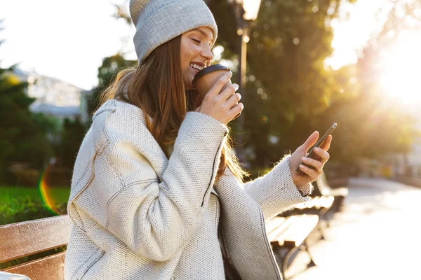 秋のコートとベンチ外に座って コーヒーを飲みながら 携帯電話を使用しての帽子に身を包んだ美しい若い女性 — ストック写真