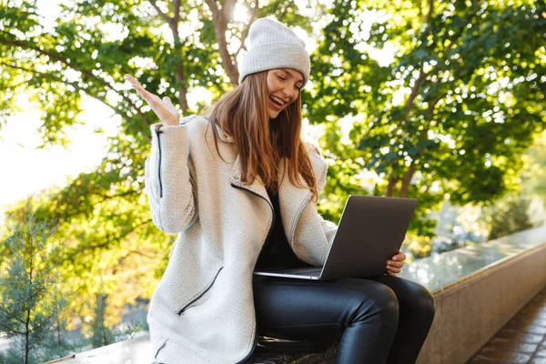 秋のコートと屋外に座って ラップトップ コンピューターを使用して 成功を祝っての帽子に身を包んだ美しい若い女性 — ストック写真