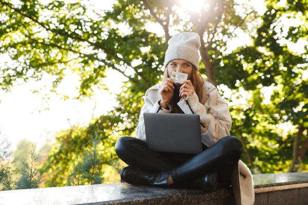 幸せな若い女を着た秋コートと帽子は屋外 ラップトップ コンピューターを使用して プラスチック製のクレジット カードを見せて — ストック写真