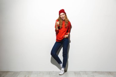 Tam uzunlukta başparmak gösterilmesini hoodie ayakta beyaz arka plan üzerinde giyen bir mutlu genç kadın portresi