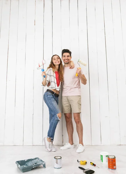 Full length photo of loving couple man and woman painting white wall and making renovation using paint rollers