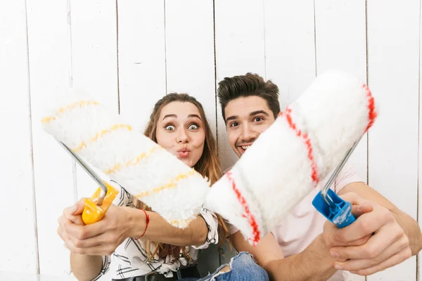 Image of young couple man and woman holding paint rollers and standing over white wall while painting and making renovation