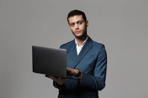 分離ラップトップを使用して 灰色の背景の上に立っているスーツを着て自信を持って若い実業家の肖像画 — ストック写真
