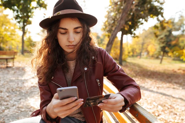 クレジット カードで携帯電話を使用して 公園のベンチに座ってきれいな若い女の子 — ストック写真
