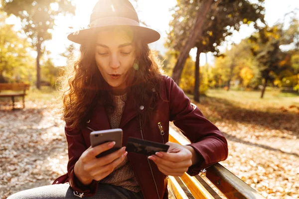 クレジット カードを保持している携帯電話を使用して公園のベンチに座っているかわいい美人の画像 — ストック写真