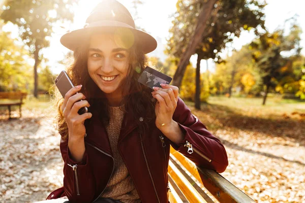 クレジット カードを保持している携帯電話を使用して公園のベンチに座っているかわいい美人の画像 — ストック写真
