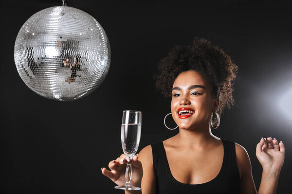 Beautiful african woman standing with silver disco ball isolated over black background, holding glass of champagne