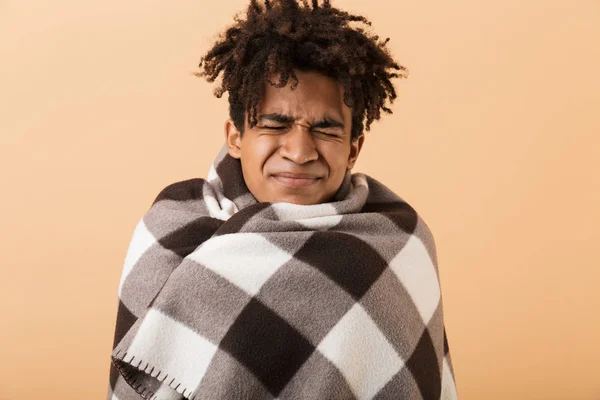 Portrait of frozen african american guy frowning while covering in blanket isolated over beige background