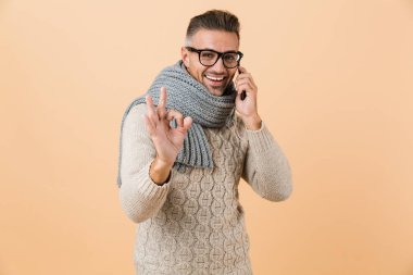 Gülümseyen bir adam kazak ve bej renkli arka plan üzerinde izole ayakta eşarp ok gösterilen cep telefonda konuşurken giymiş Eğer portre