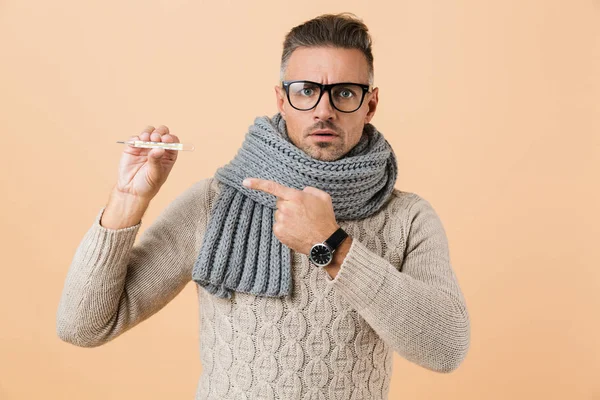 Portrait of a sick man dressed in sweater and scarf standing isolated over beige background, showing thermometer