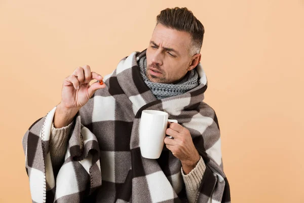 Close up portrait of a confused man wrapped in a blanket standing isolated over beige background, showing pill capsule