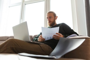 Belgeleri analiz etme evde çalışma, dizüstü bilgisayar, kullanan bir kanepede oturan yakışıklı genç adam