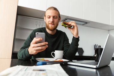 Evde dizüstü bilgisayar üzerinde çalışan, mutfak oturma, çay içme, yemek yiyor, cep telefonu tutan meşgul genç adam