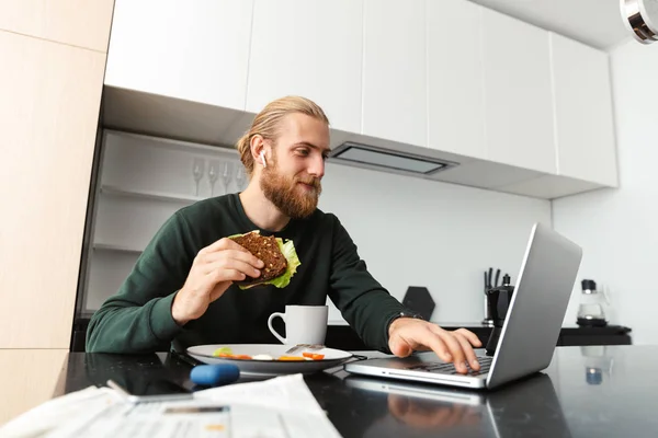Joven Ocupado Trabajando Computadora Portátil Casa Sentado Cocina Bebiendo Almorzando — Foto de Stock
