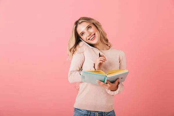 一个开朗的年轻女子穿着在粉红色背景上被孤立的毛衣的肖像 在手机上交谈时在日记中写笔记 — 图库照片