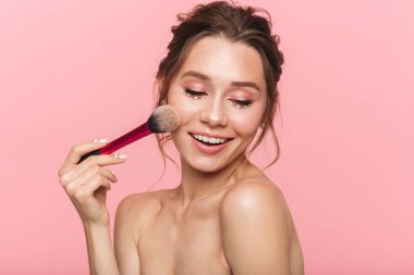 Beyaz inci giymiş pembe arka plan üzerinde izole bir fırça ile makyaj uygulamak çekici bir genç üstsüz kadın güzellik portresi