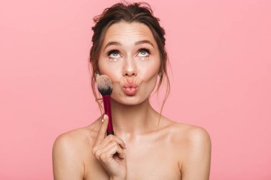 Beyaz inci giymiş pembe arka plan üzerinde izole bir fırça ile makyaj uygulamak çekici bir genç üstsüz kadın güzellik portresi